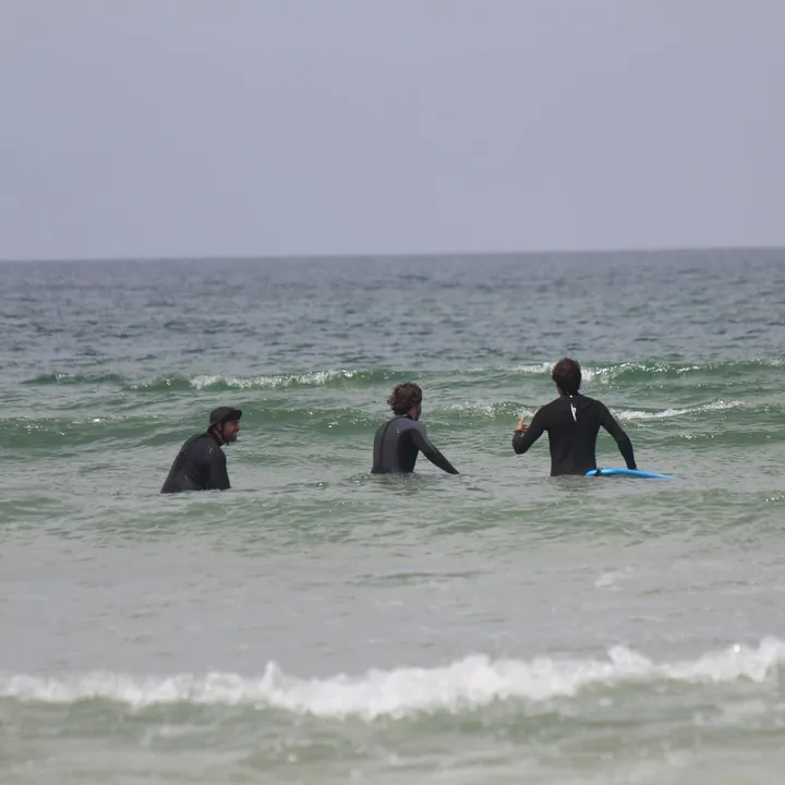 Three surfers in wetsuits waist-deep in green water, waiting side by side as a small wave breaks past them.