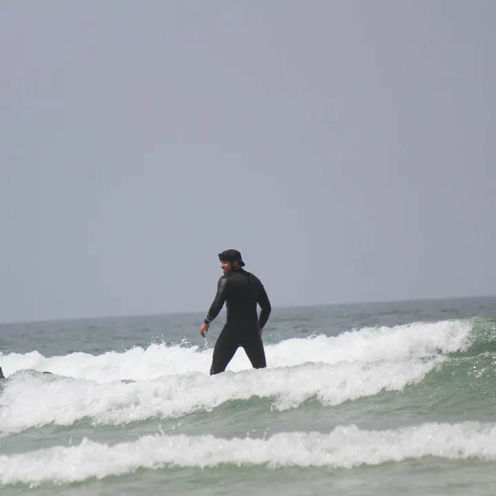A surfer in a black wetsuit and cap standing waist-deep in the white water, looking back over his shoulder and grinning.