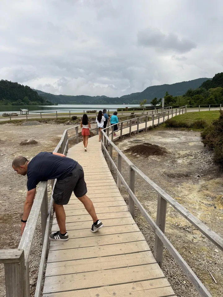 A wooden boardwalk winding across bare steaming ground beside a lake at Furnas, walkers ahead of him and Ismael leaning on the rail in the foreground.