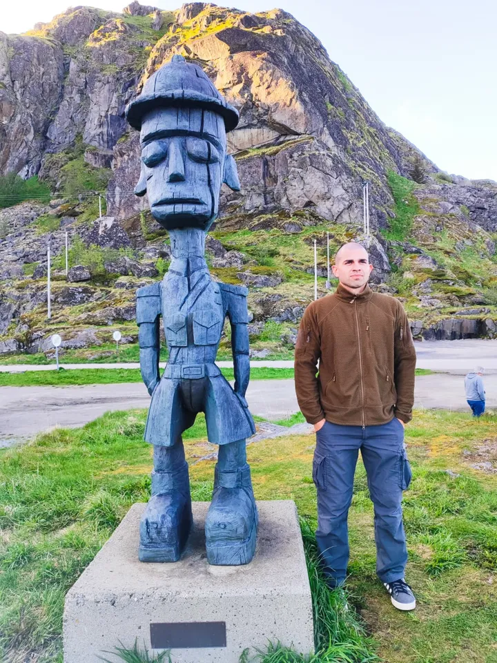 A tall carved wooden figure in a fisherman's hat on a plinth, Ismael standing beside it below a steep green and grey mountain in Lofoten.